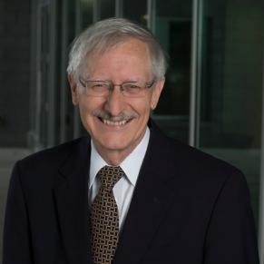 A man in a dark jacket and tie with eyeglasses smiles at the camera.