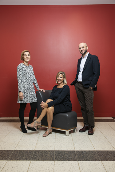 A coed group of 3 adults wearing professional clothing and posing for a photo in front of a red wall.