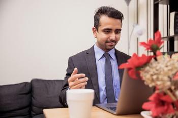 Man wearing a suit sitting at a desk, speaking in the direction of a laptop screen.